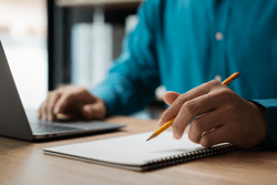 A man is writing on a notebook with a pencil. He is focused on his work and he is in a serious mood. The notebook is placed on a wooden desk, and there is a laptop nearby