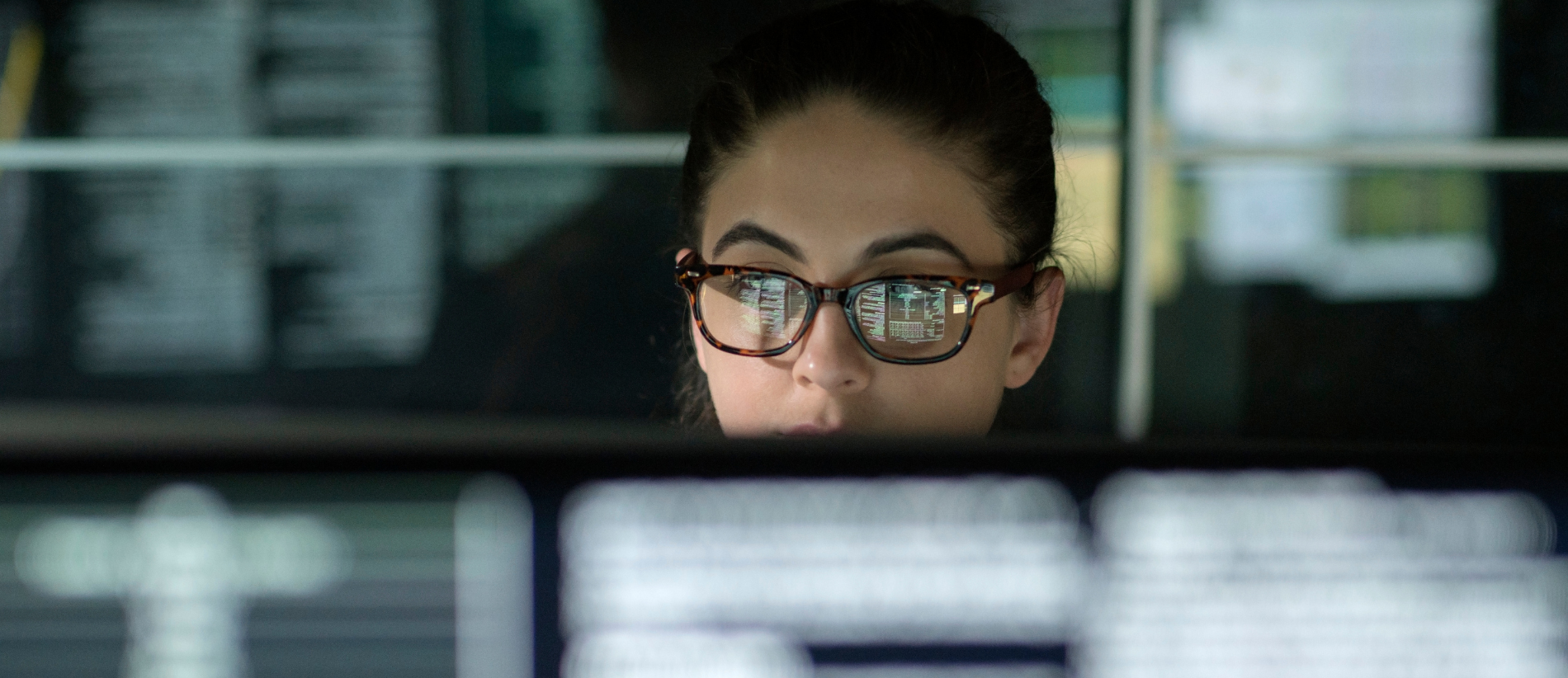 A young woman is surrounded by monitors & their reflections displaying scrolling text & data.