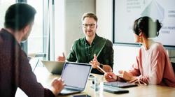 A focused team leader is addressing colleagues in a meeting, with a presentation displayed in the background, capturing a moment of active corporate engagement.