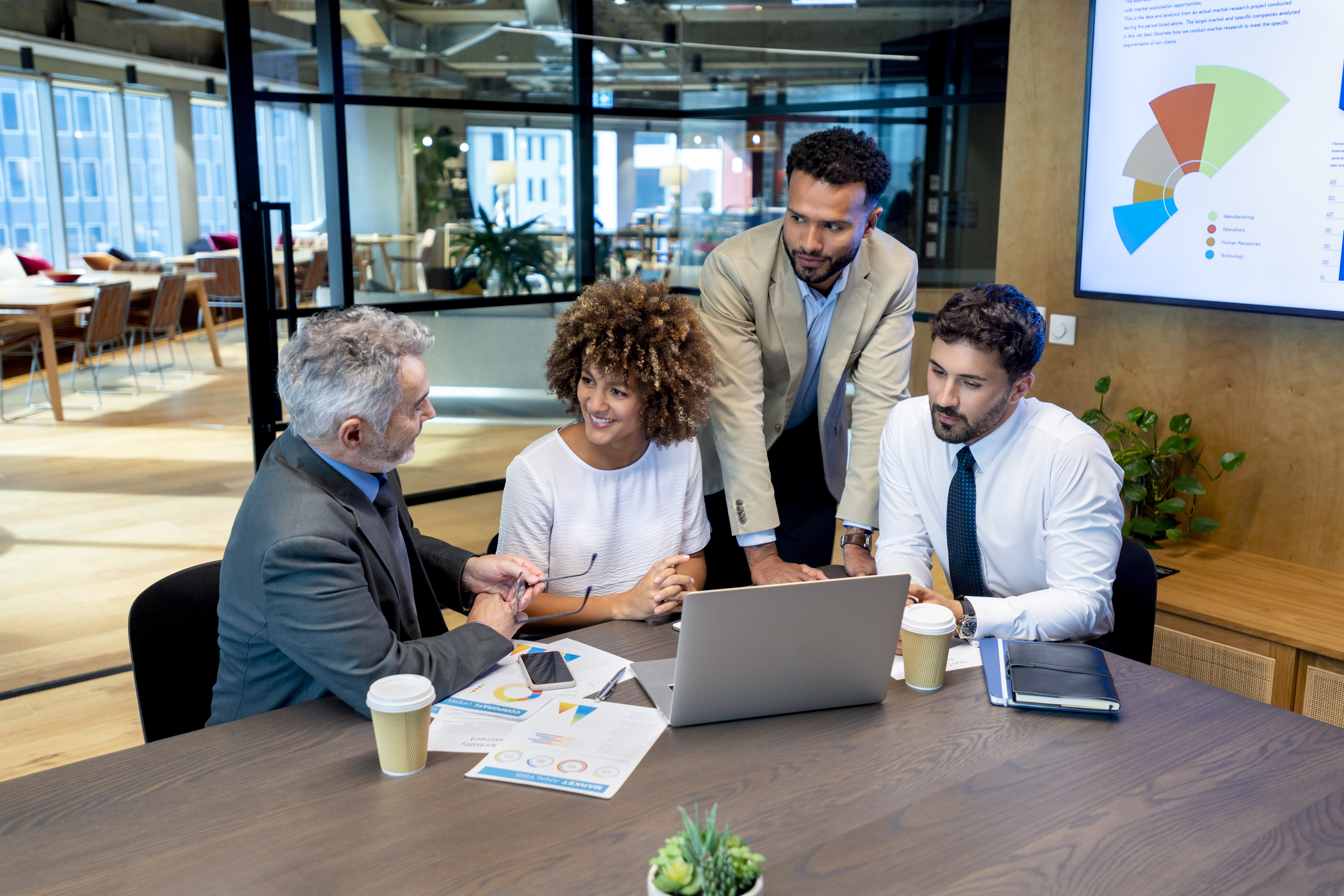 Four business people working on a laptop computer in an office. There is paperwork on the table with financial figures, graphs and charts. There is also a tv in the background with charts and graphs