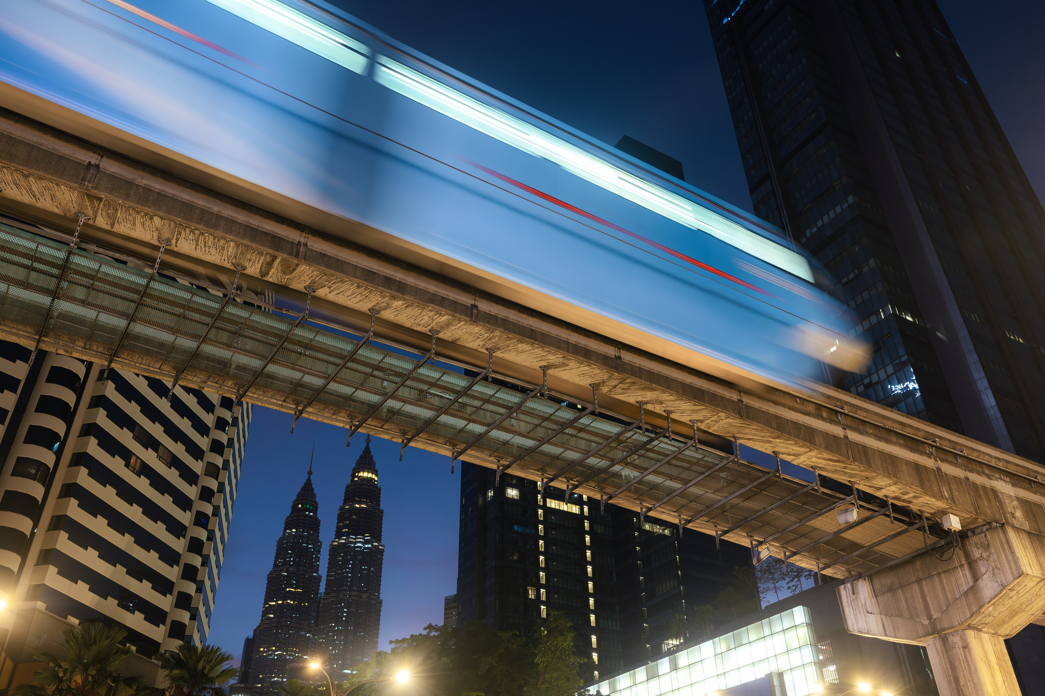 Elevated monorail between tall buildings and Petronas Towers in background in Kuala Lumpur.