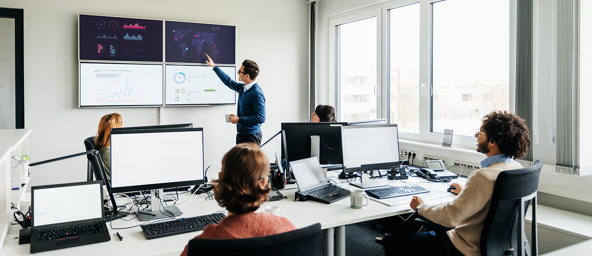 A business office with a number of employees working at computers while listening to a colleague talking and reviewing some data on wall mounted monitors.