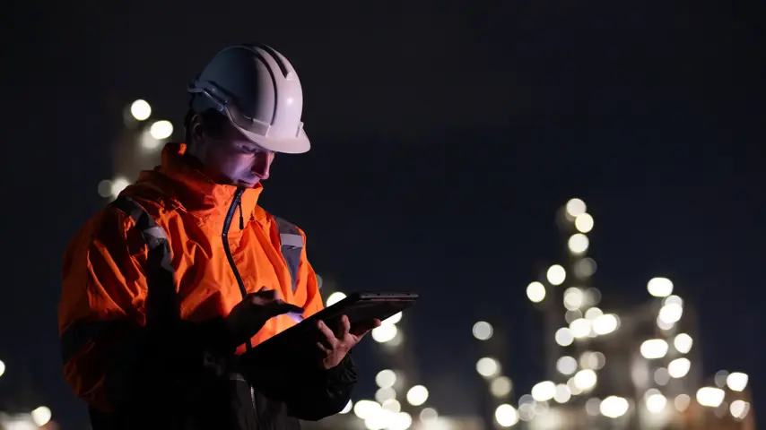 A night shift engineer in a hard hat and reflective jacket is focused on a tablet with the illuminated industrial plant in the background.