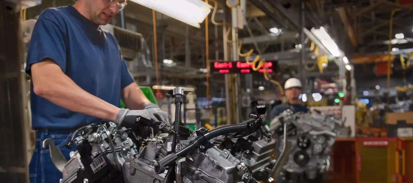 A tuo assembly plant worker adds components to an internal combustion engine before it is installed in the chasis of a car.