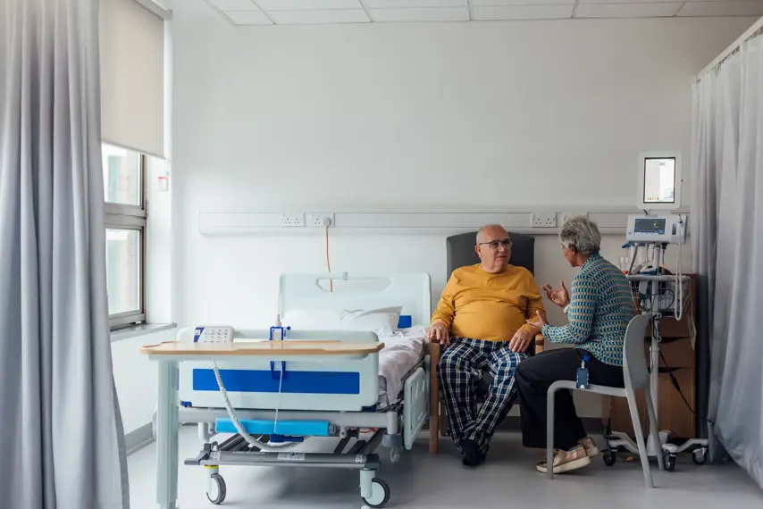 A senior male patient with cancer staying on a ward in a hospital in Newcastle upon Tyne, North East England. He is sitting on a chair next to his hospital bed and talking to a female consultant, who is sitting next to him on a chair. They are discussing his treatment options and how they are going to progress with his condition.