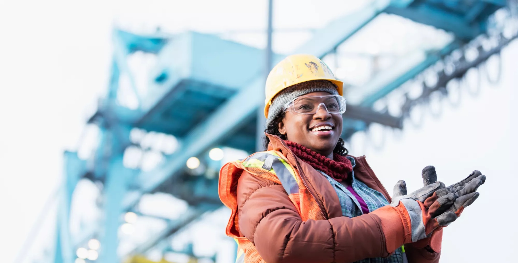 A mid adult African-American woman in her 30s wearing a hard hat, safety vest and safety goggles, a dock worker working at a shipping port. A gantry crane is out of focus in the background.