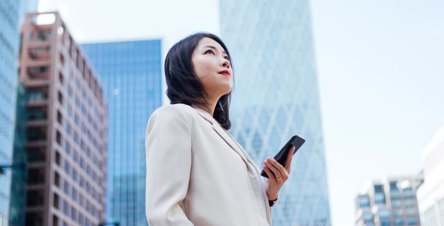 Low angle view of beautiful young Asian woman using smartphone while standing in front of commercial buildings in financial district. Young professional looking for a new career opportunity.