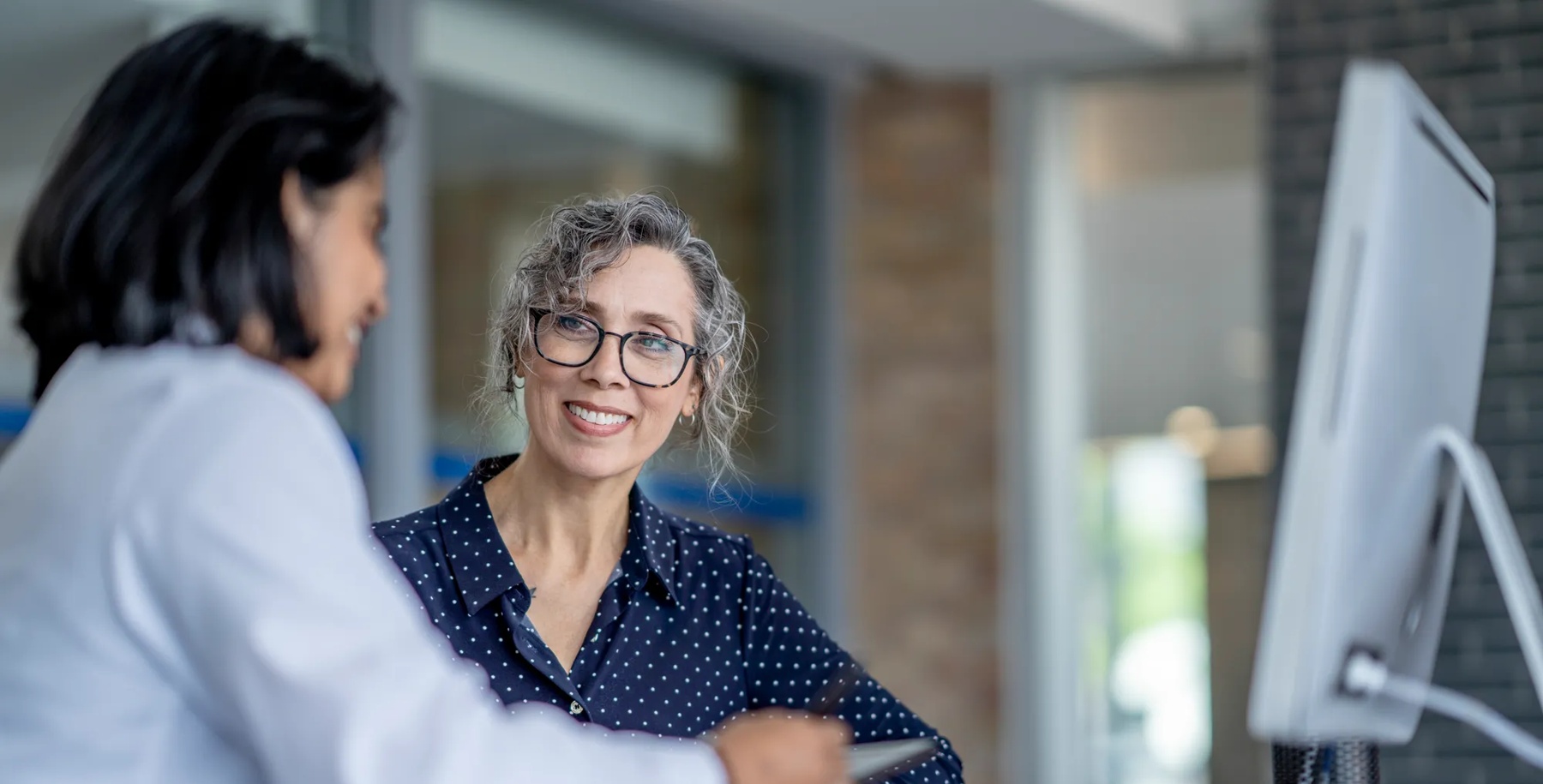 A middle aged woman sits with her female doctor of Middle Eastern decent as they review her medical concerns.  She is dressed semi-casually and sharing with the doctor her recent discomfort.  The doctor is wearing a white lab coat and taking notes on her tablet as the two talk.