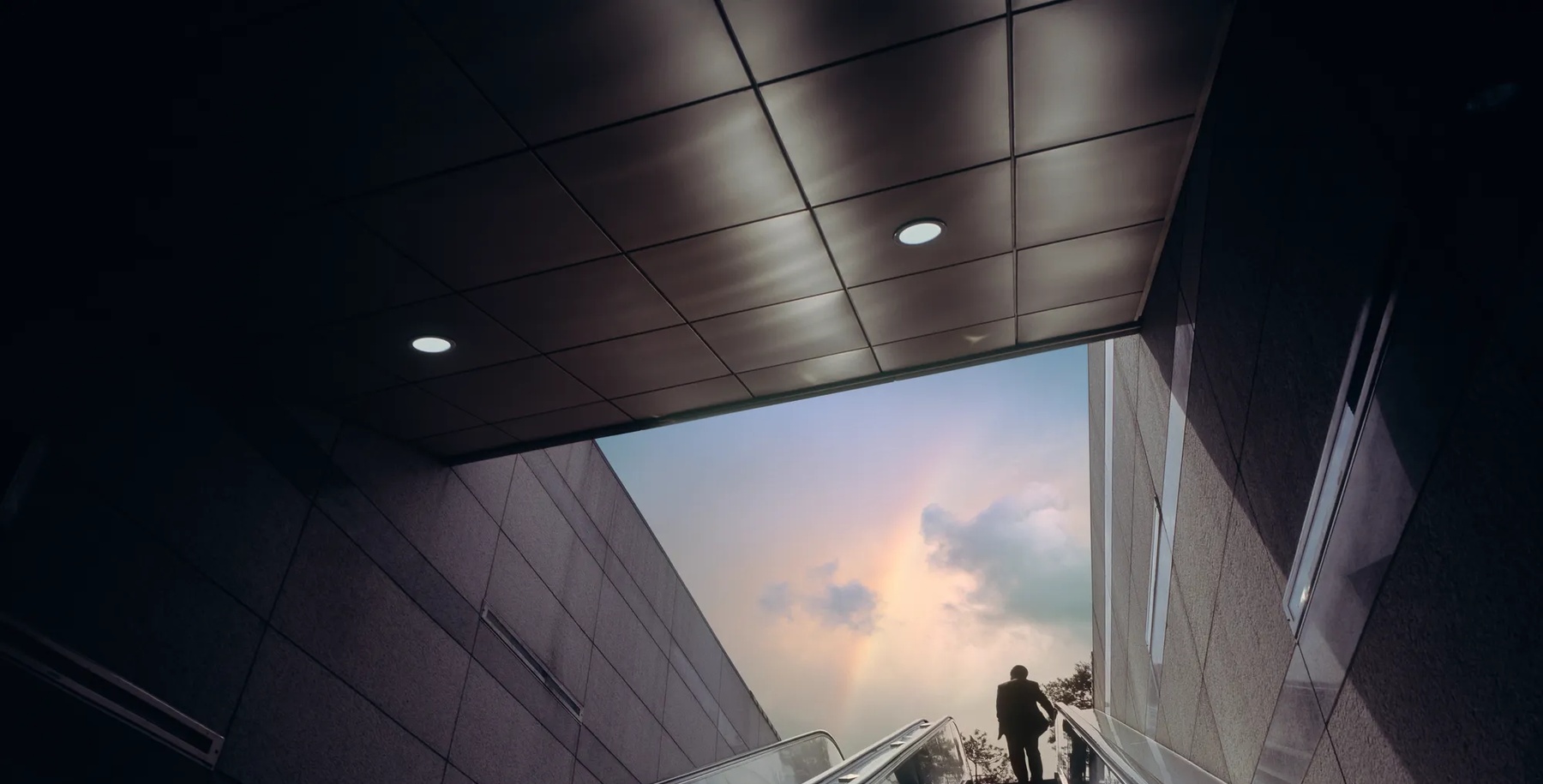 Escalator of subway station with businessman moving towards dramatic sky with rainbow.