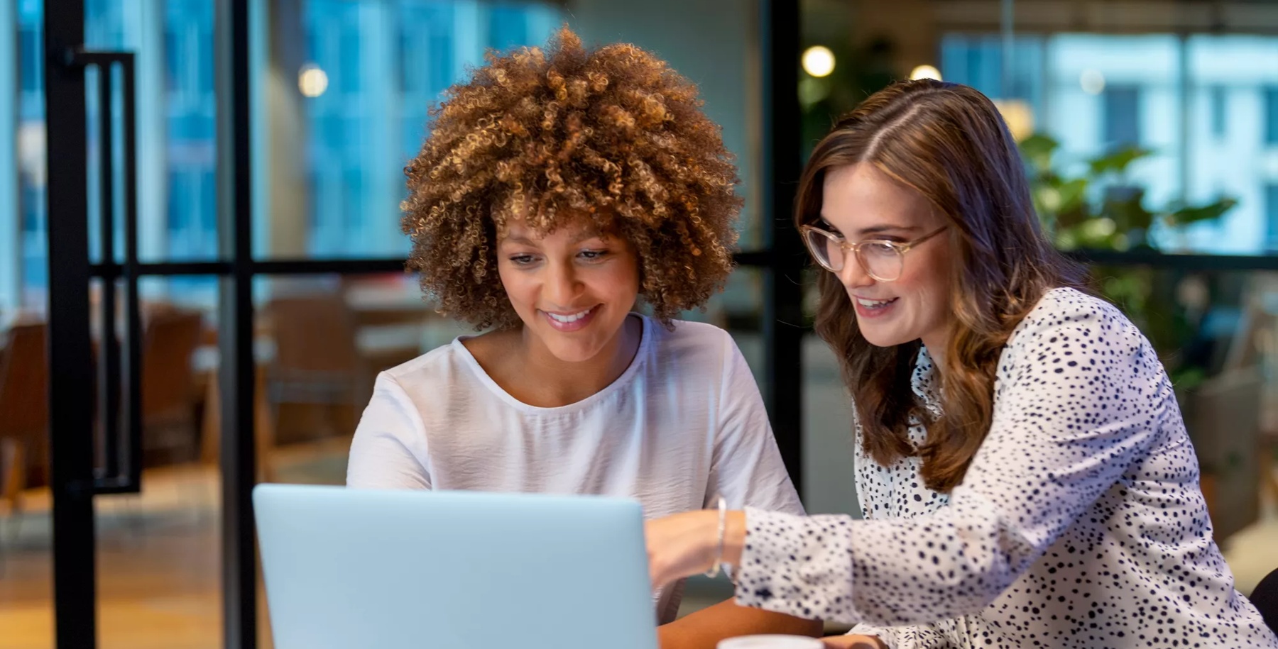 Business colleagues working together on a laptop. They are both young business people dressed in business attire in a modern office. Could be an interview, training or consultant working with a client. She is listening, talking and smiling. There is paperwork, coffee and technology on the table. Mixed ethnic group.