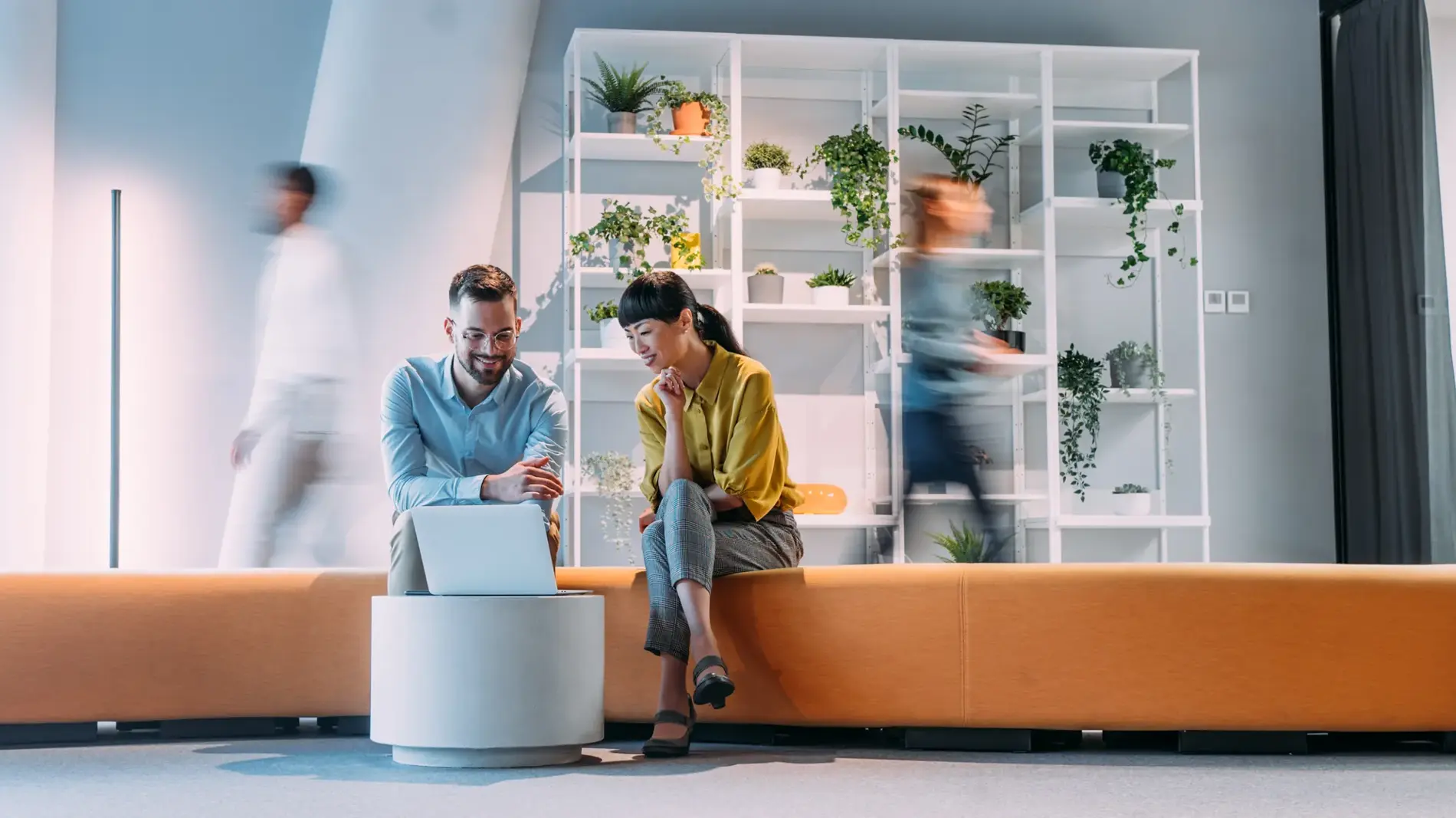 Shot of business people having a meeting in modern office. Blurred motion of energetic businesspeople on the move and project team members discussing ideas in a office lobby. Businesspeople walking in blurred motion around their colleagues. Business persons sitting on a sofa and using laptop while their colleagues are walking in blurred motion. Creative business team on meeting in the office.