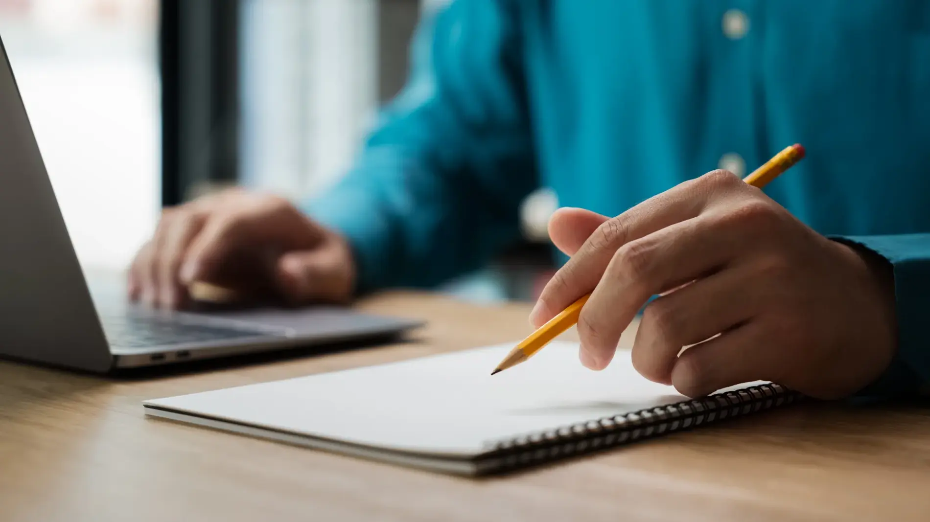 A man is writing on a notebook with a pencil. He is focused on his work and he is in a serious mood. The notebook is placed on a wooden desk, and there is a laptop nearby