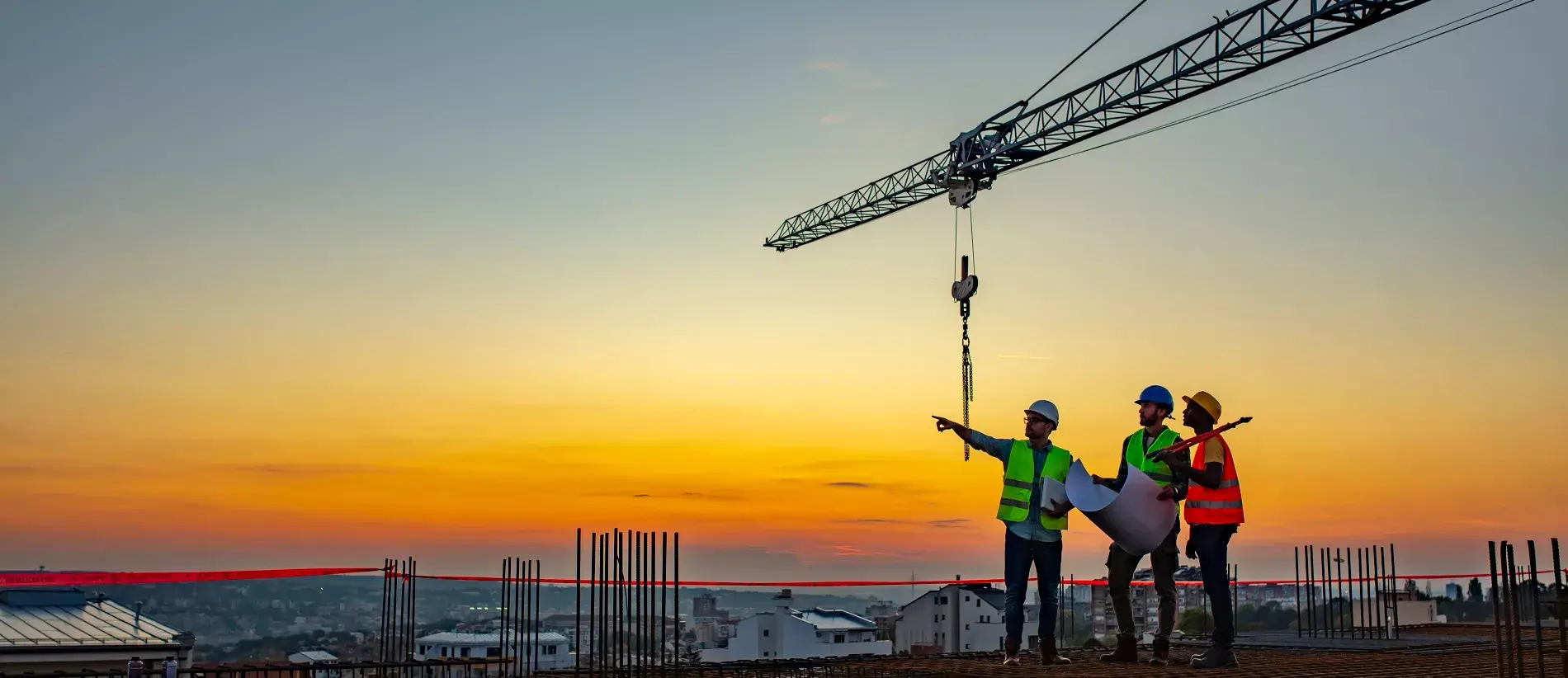 Three Multi-Ethnic construction workers in uniform standing at construction site with crane in background, discussing building plans while holding blueprint at sunset under the tower crane.