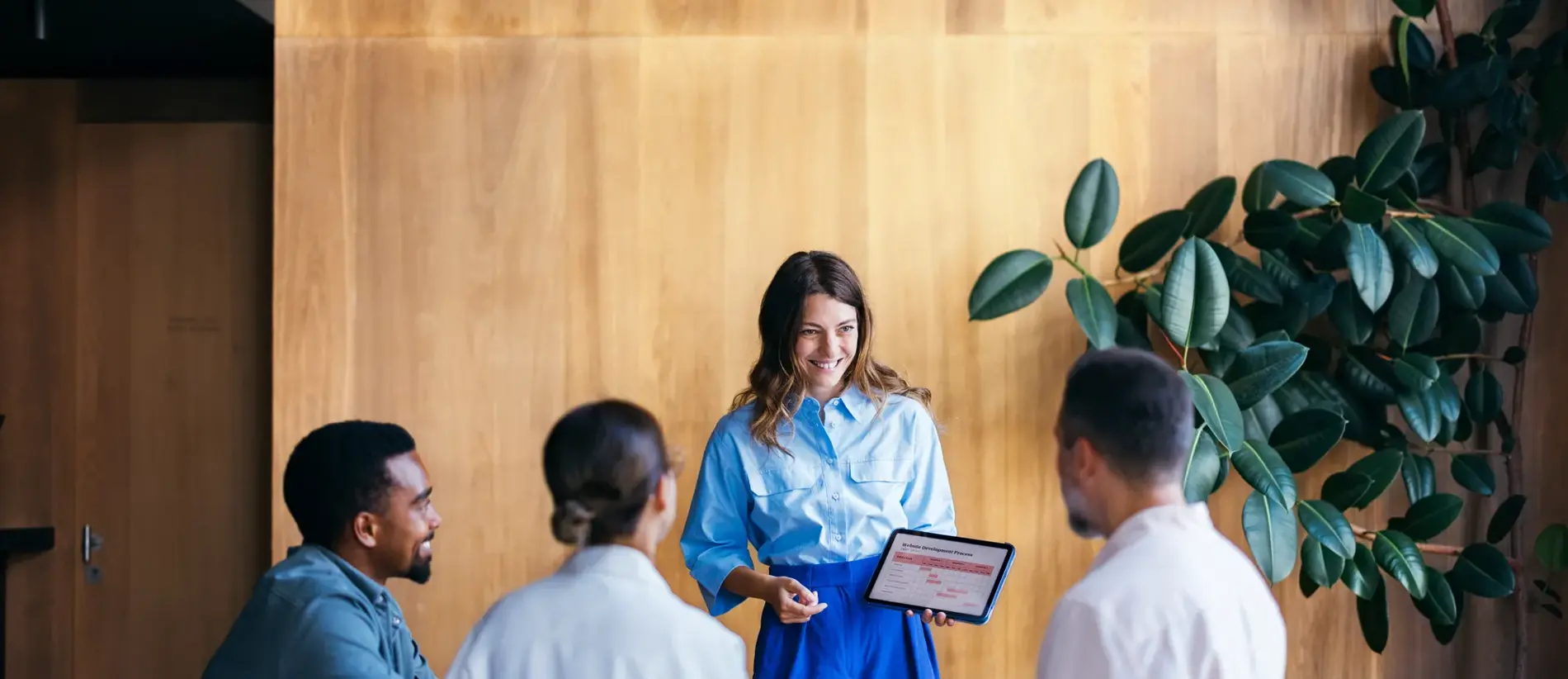 A team collaborates in a modern office room, engaged in discussion as one presents information using a tablet and documents. The setting features a wooden wall and green plant for a professional ambiance.