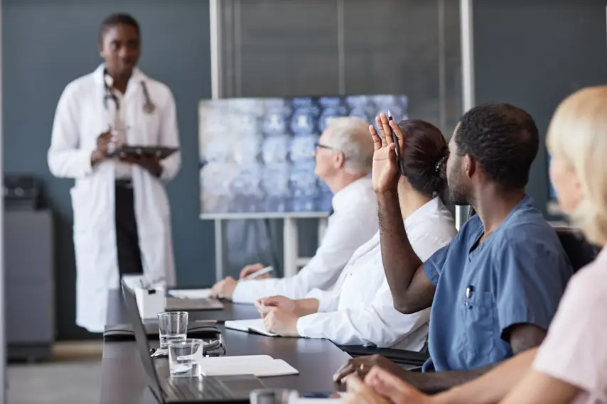 Side view of African American male clinician raising hand asking colleague follow up question after presentation in group seminar while sitting at table in conference room, copy space