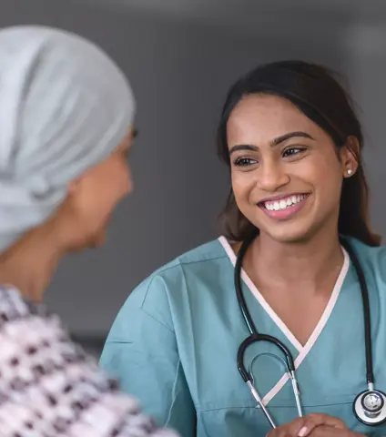 A mixed-race female doctor is meeting with a female patient with cancer. The two individuals are seated next to each other at a table.The physician is facing the camera. She is smiling warmly at the patient. The patient is wearing a bandana to hide her hair loss from chemotherapy treatment.