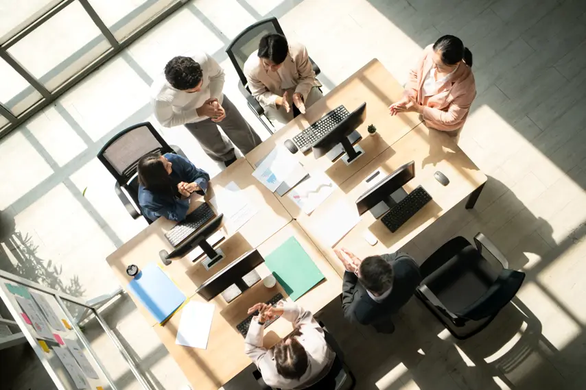 Business team standing around a conference table during meetings, discussing charts and reports under natural light, and congratulating each other on achievements with applause