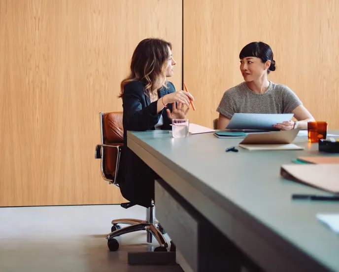 Two businesswomen engaged in a discussion at a modern office table. They are collaborating on a project with documents and laptops.