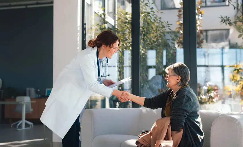 A smiling female doctor shakes hands with a senior woman in a bright hospital waiting area, conveying trust, compassionate care, patient-doctor communication and positive health support.