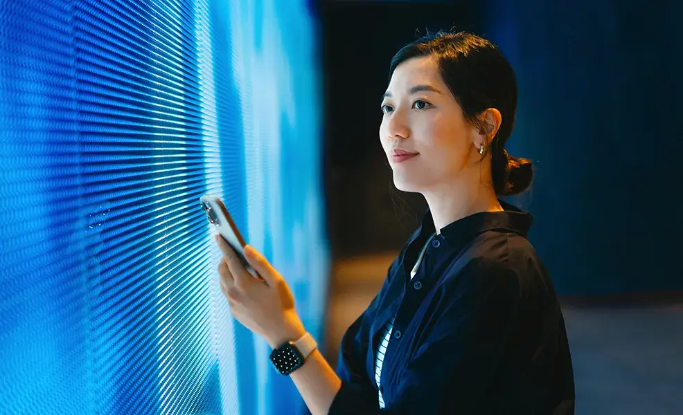 Confident young Asian woman using smartphone against blue coloured illuminated LED digital display screen in the dark. Connecting to the future. Futuristic. Artificial intelligence. Innovation, lifestyle and technology. Smart banking with technology